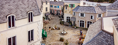 European-style shopping village with stone walkways, boutique storefronts, and central fountain.